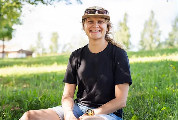 women sitting in green grass, with jean shorts and black tshirt on. Smiling wearing a hat